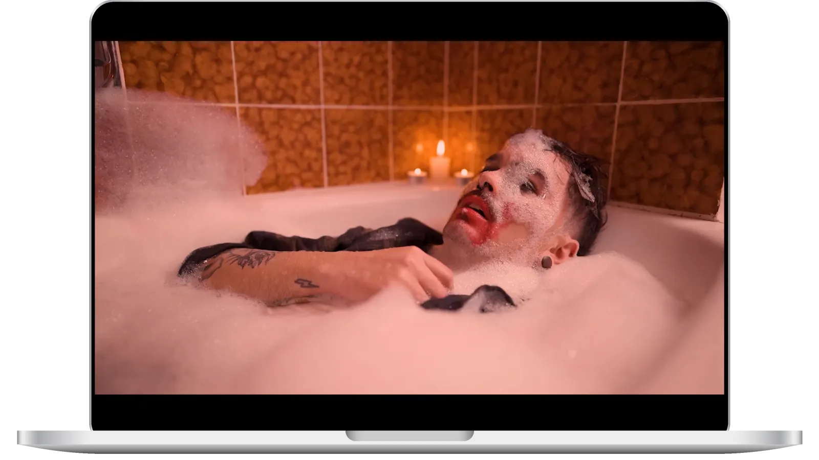 Kinematografische Szene aus dem Musikvideo "Fading Away" von Venom Breath: Ein tätowierter Mann mit Kunstblut am Mund liegt bekleidet in einer schaumgefüllten Badewanne. Im Hintergrund brennen Kerzen und sorgen für ein düster-atmosphärisches, warmes Licht.